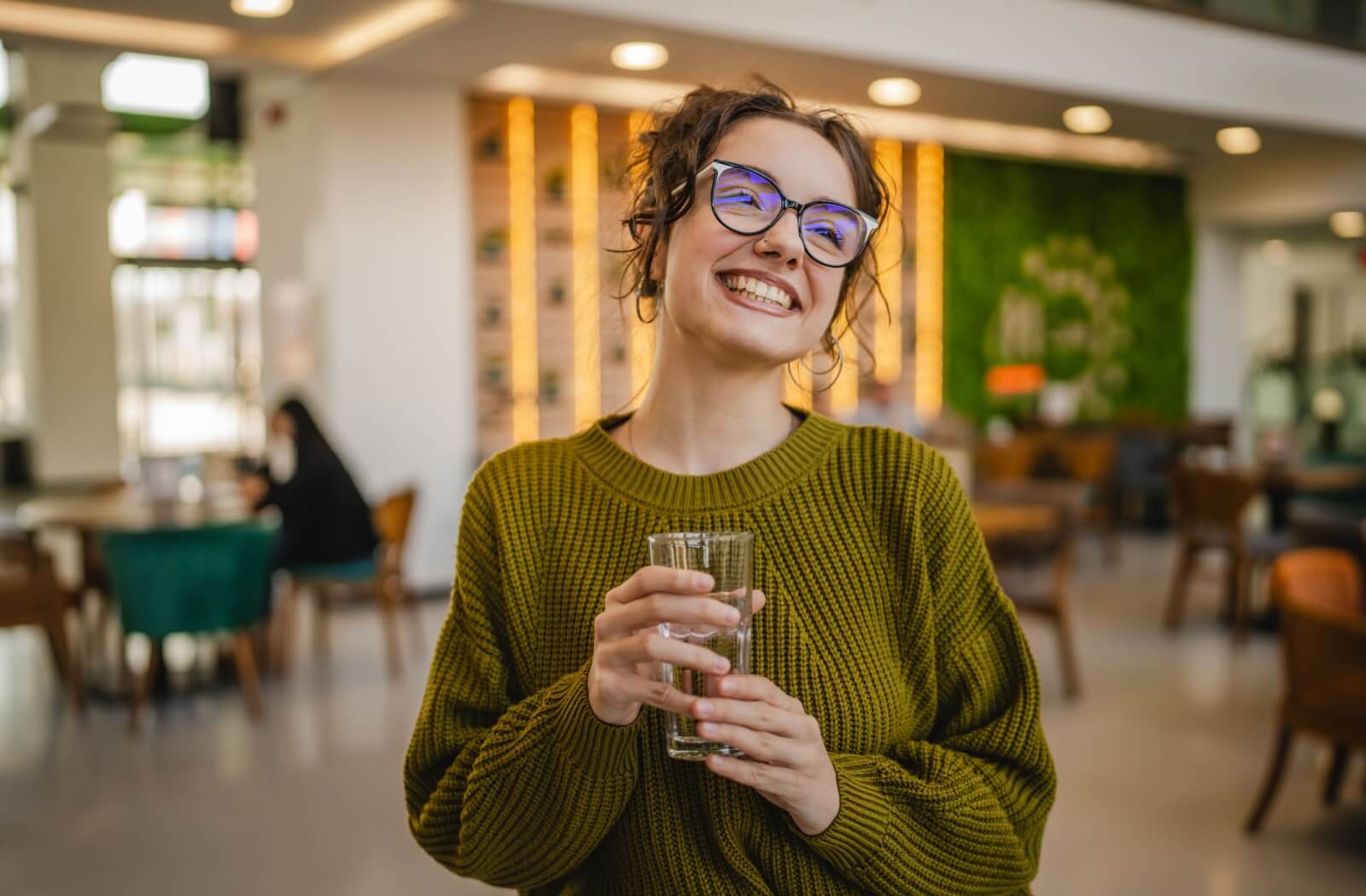 A happy person wearing stylish progressive eyeglasses while holding a glass of water, showcasing the natural visual transition and comfort of multifocal lenses.