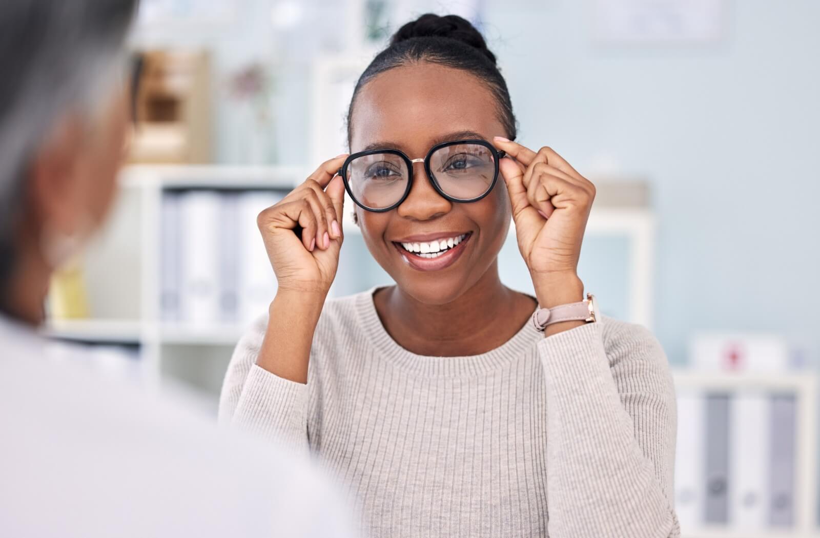 A smiling adult trying on a pair of large, round black eyeglass frames during an eye appointment.
